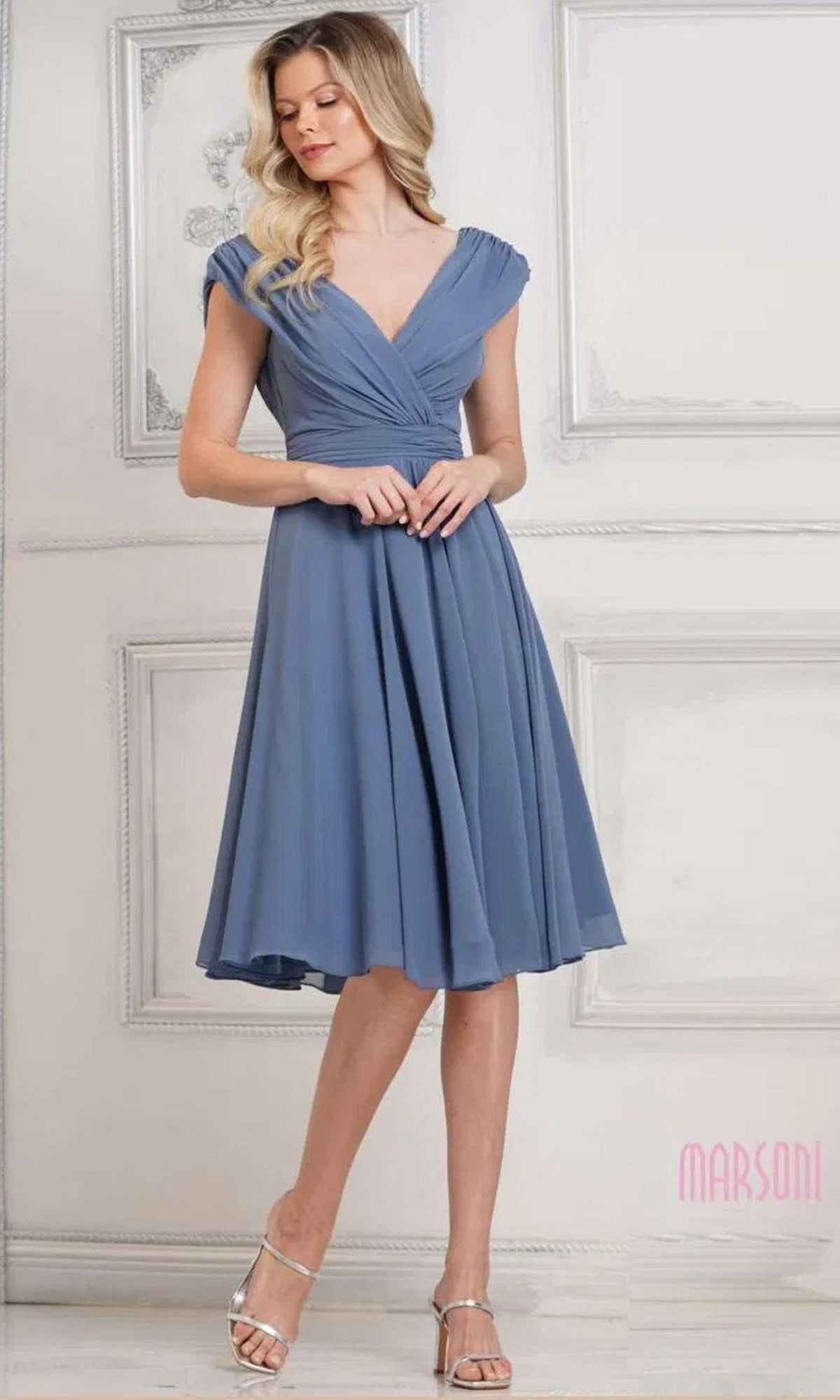 A woman wearing the Marsoni M251S short semi-formal dress in slate blue with a v-neckline and cap sleeves, paired with silver heels, stands before a decorative white wall.